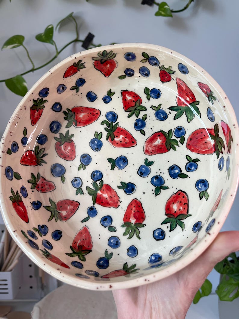 Op de afbeelding: Een keramische kom met een handgeschilderd ontwerp van aardbeien en bosbessen. De kom is wit met een roze rand en het fruit is rood en blauw. De kom heeft een diameter van ongeveer 20 cm.