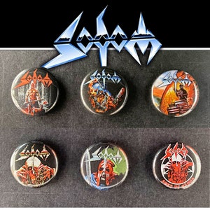 Könnte beinhalten: Sechs runde Metallpins mit schwarzem Hintergrund und einem Band mit dem Wort "SODOM" in weißen Buchstaben. Jeder Pin zeigt ein anderes Bild aus der Albumkunst des Bands.