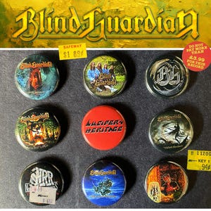 Puede incluir: Una colección de nueve botones redondos de la banda Blind Guardian. Los botones presentan varios diseños, incluyendo logotipos de la banda, arte de álbumes y el texto "Lucifer's Heritage". Algunos botones tienen etiquetas de precio. El fondo es una superficie oscura.
