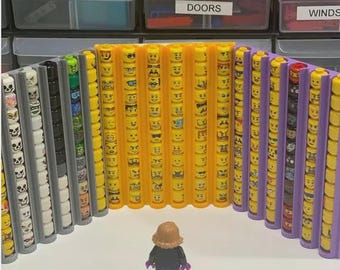 以下が含まれることがあります： さまざまな顔の表情を持つ黄色のレゴヘッドのコレクション。オレンジ、グレー、紫のホルダーに縦に並んでいます。レゴのフィギュアがディスプレイの前に立っています。背景には透明な収納ボックスがあります。