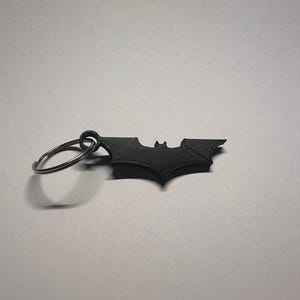 Könnte beinhalten: Ein schwarzer Metall-Schlüsselanhänger mit dem Fledermaussymbol. Der Schlüsselanhänger hat einen kreisförmigen Ring zum Befestigen von Schlüsseln und eine detaillierte Fledermaus-Silhouette. Ein erkennbares Emblem.