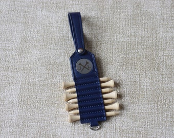 May include: A navy blue leather golf tee holder with a silver monogrammed disc featuring the initials "J.L." and crossed axes. The holder has a loop for attaching to a golf bag and contains six wooden golf tees.