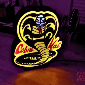 May include: Illuminated Cobra Kai logo sign with a black background and yellow and red accents. The sign features a cobra head design with the words "Cobra Kai" in red. The sign is lit up, creating a glowing effect.