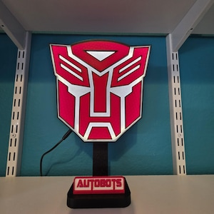 Könnte beinhalten: Ein rotes und weißes Transformers Autobots Logo-Licht. Das Logo ist auf einem schwarzen Ständer mit dem Wort "AUTOBOTS" auf der Basis montiert. Das Licht ist mit einem schwarzen Kabel angeschlossen.