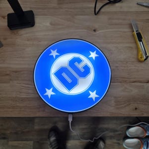 May include: A circular, illuminated sign featuring the blue and white DC logo in the centre, surrounded by four white stars on a vibrant blue background. The sign is powered by a white USB cable connected to the bottom edge of the circular frame.