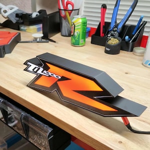 Puede incluir: Gráfico negro y naranja con el texto "Z1000 R" sobre una superficie de madera. El gráfico tiene un diseño geométrico con una "R" estilizada en naranja. Un cable negro se extiende desde el gráfico.