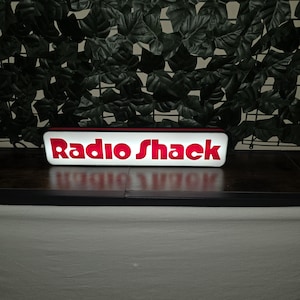 May include: A rectangular, illuminated sign featuring the red text RadioShack on a white background. The sign has a black frame and is positioned horizontally, emitting a bright glow from its front panel, displaying the brand name clearly.