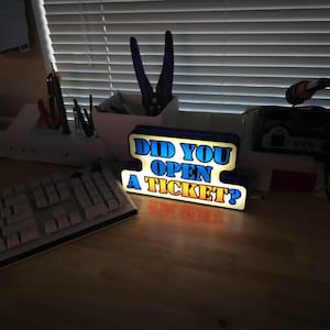 May include: A lighted sign with the text "DID YOU OPEN A TICKET?" in blue and yellow. The sign is on a wooden desk, with a keyboard and tools in the background.