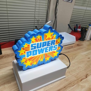 May include: A blue and yellow "Super Powers" sign with a starburst design. The sign is mounted on a white rectangular base. The text is in a bold, comic book style font.