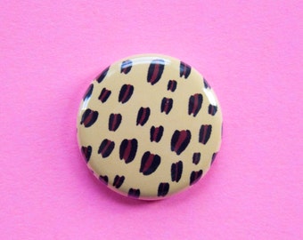 Puede incluir: Un botón redondo con un diseño de estampado de leopardo. El botón es beige con manchas negras y burdeos.