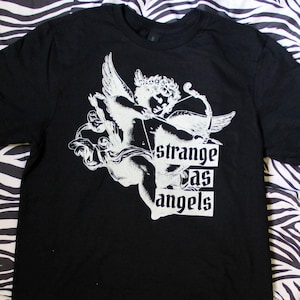Op de afbeelding: Zwart T-shirt met een witte afbeelding van een cherubijn die een boog en pijl vasthoudt. De tekst "strange as angels" is onder de cherubijn gedrukt.