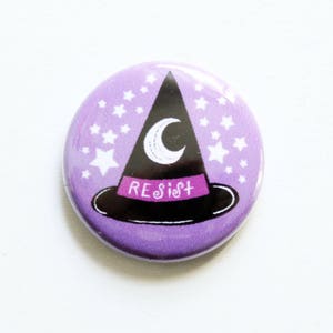 May include: Purple button with a black witch hat with a crescent moon and the word "Resist" on the hat brim. The hat is surrounded by white stars.