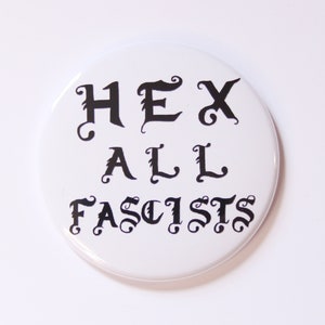 May include: White round button with black text that reads "HEX ALL FASCISTS" in a stylized font.