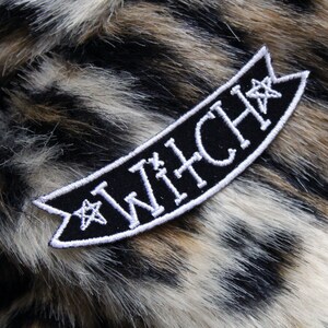 May include: Black fabric patch with white embroidered text that reads "WITCH" with a star on either side of the text. The patch is on a brown and white faux fur background.