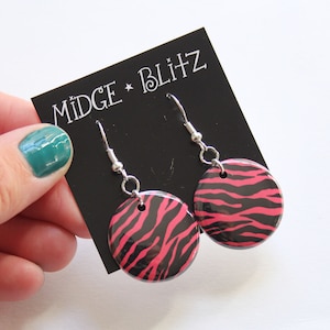 Puede incluir: Pendientes redondos con un estampado de cebra en negro y rosa intenso. Los pendientes se muestran sobre una tarjeta negra con el texto "MIDGE * BLITZ".