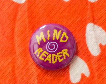 Könnte beinhalten: Lilafarbener Knopf mit gelbem Text, der "MIND READER" mit einem Wirbelmuster in der Mitte lautet.