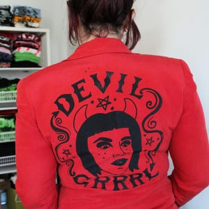 Könnte beinhalten: Roter Blazer mit schwarzer Grafik auf dem Rücken. Die Grafik zeigt ein Teufelsgesicht mit dem Text "DEVIL GRRR" in einer stilisierten Schriftart. Der Blazer hat eine einfarbige rote Farbe.