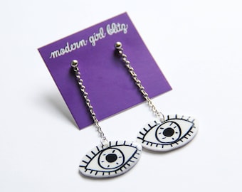 Puede incluir: Un par de pendientes de plata con colgantes blancos en forma de ojo. Los pendientes están en una tarjeta morada con el texto "modern girl blitz".