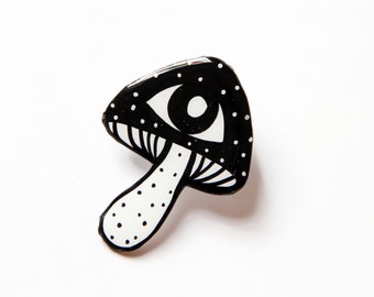 Puede incluir: Pin de esmalte negro y blanco de un hongo con un ojo en el sombrero. El hongo tiene un tallo blanco con lunares negros.