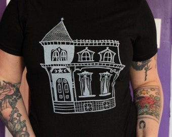 May include: Black t-shirt featuring a white graphic of a spooky house with a star and crescent moon above the door.