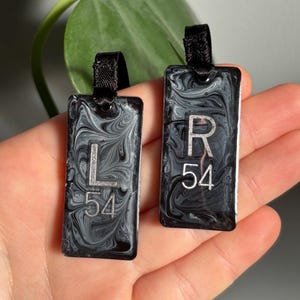 May include: Two black and white marbled rectangular tags with black ribbon loops. The left tag has a silver 'L' and '54' printed on it. The right tag has a silver 'R' and '54' printed on it.