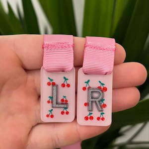 May include: Two pink rectangular tags, each with a silver letter, "L" and "R". The tags are decorated with red cherry illustrations and have a pink ribbon at the top. These tags are likely for labeling items.