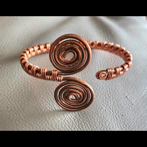May include: Copper wire cuff bracelet with two spiral designs. The bracelet is made of coiled copper wire, with a spiral design at the center and another at the end. The bracelet is on a light gray surface.