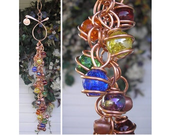 May include: A copper wire wind chime with a spiral design and colorful glass beads. The beads are in a rainbow pattern, with red, orange, yellow, green, blue, indigo, and violet beads. The wind chime is hanging from a copper wire loop.