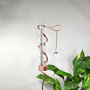 May include: Copper rain gauge with a crystal prism hanging from it. The gauge is attached to a copper wire that is coiled into a spiral shape. The gauge is placed in front of a green plant with yellow variegation.