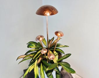 May include: A set of five copper-colored mushroom-shaped garden stakes with hammered textures on the caps, inserted into a potted plant with variegated green and yellow leaves. The stakes feature thin stems and rounded tops.