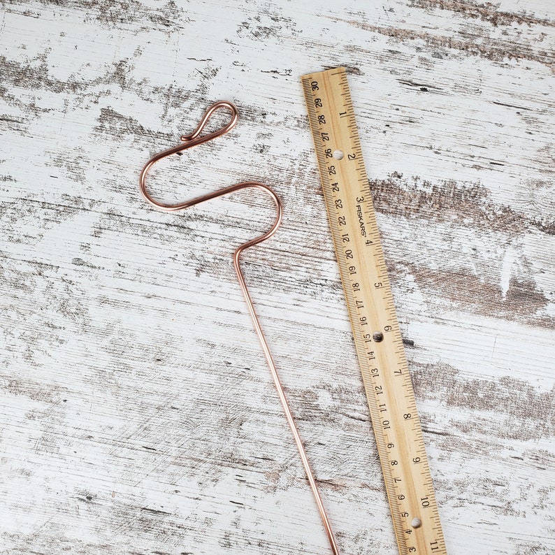 Puede incluir: Un colgador de plantas de alambre de cobre con un dise&ntilde;o &uacute;nico en forma de S. El colgador mide aproximadamente 18 cm de largo y se muestra junto a una regla para la escala.