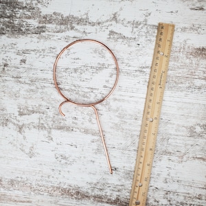 May include: A copper-colored metal plant hanger with a circular top and a long, thin stem. The hanger is designed to hold a small plant pot.