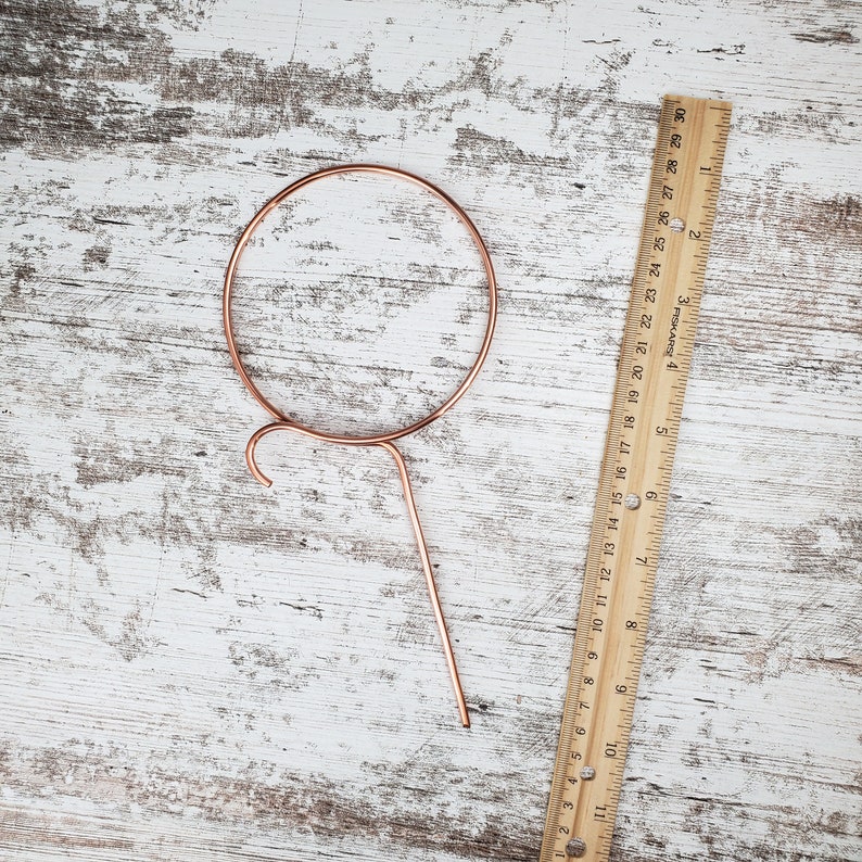 May include: A copper-colored metal plant hanger with a circular top and a long, thin stem. The hanger is designed to hold a small plant pot.