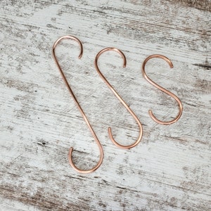 May include: Three copper-colored S-shaped hooks of different sizes. The hooks are made of metal and have a smooth, shiny finish.