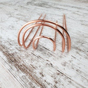 May include: A set of three copper hair combs in the shape of an arch. The combs are arranged in a graduated size, with the largest arch at the bottom and the smallest arch at the top.