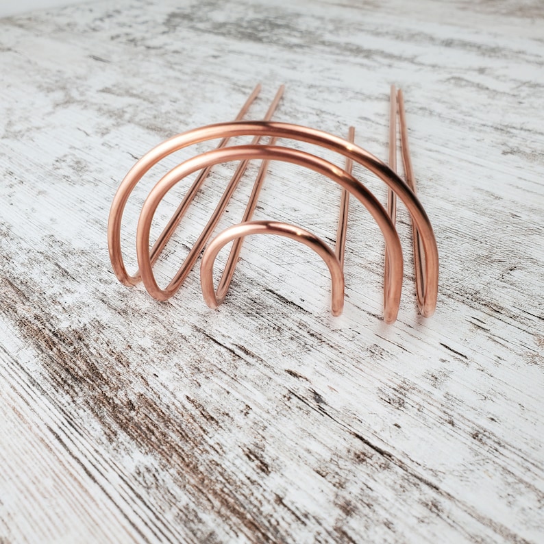 May include: A set of three copper hair combs in the shape of an arch. The combs are arranged in a graduated size, with the largest arch at the bottom and the smallest arch at the top.