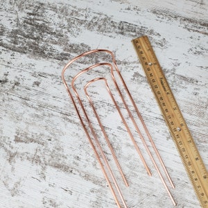 May include: Set of three copper wire hoops, nested together, on a white wooden surface. The hoops are different sizes and are all open at the top.