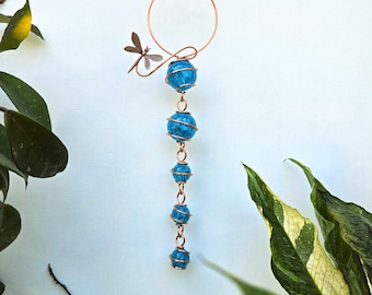 May include: A copper-colored wind chime featuring a dragonfly accent and a series of blue, spherical beads. The beads are wrapped in copper wire, and the chime hangs from a circular copper ring. The design is set against a light blue background.