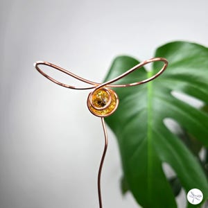 May include: A copper wire sculpture featuring a swirling design with a central, circular element containing a golden-yellow, gem-like material. The sculpture has a long stem and a looped top, with a green leaf in the background.