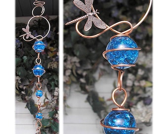 May include: Copper rain chain with blue glass orbs and dragonfly accents. The rain chain features a series of blue glass spheres, each encircled by copper wire, and a decorative dragonfly. The chain is designed to hang from a gutter.