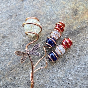 May include: A decorative garden stake featuring copper wire stems twisted into spirals. One stem holds a large crackled clear glass bead and a copper dragonfly charm. Two other stems feature alternating red, white, and blue crackled glass beads.
