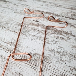 May include: Two copper wire plant hangers with a simple design. The hangers are made of a single piece of wire that is bent into a loop at the top and then into a hook at the bottom. The hangers are shown against a white wooden background.