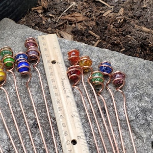 May include: A set of 15 copper wire garden stakes with glass beads in various colors, including red, orange, yellow, green, blue, and purple. The stakes are approximately 6 inches long and are arranged on a gray stone surface next to a ruler.