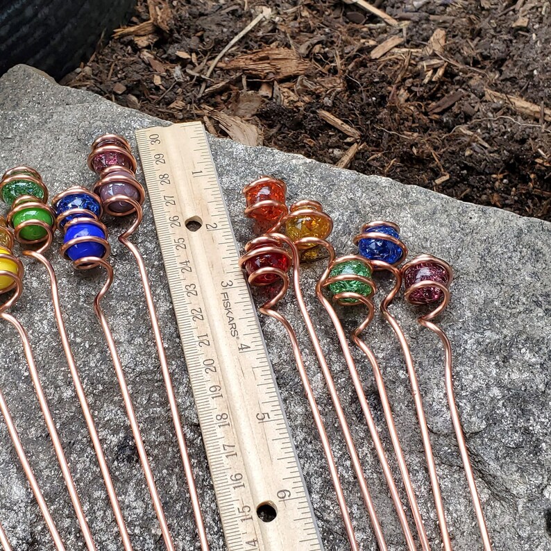 May include: A set of 15 copper wire garden stakes with glass beads in various colors, including red, orange, yellow, green, blue, and purple. The stakes are approximately 6 inches long and are arranged on a gray stone surface next to a ruler.