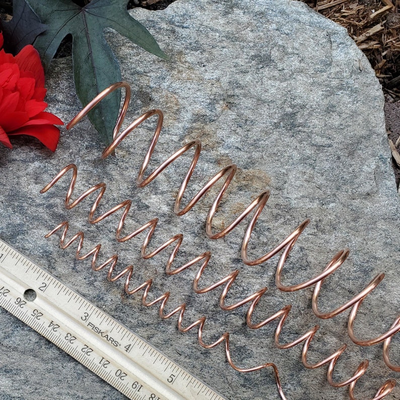 May include: Three copper wire coils of different sizes, arranged on a gray rock surface. The coils are all in a spiral shape and are made of a thin, flexible wire.