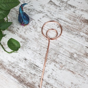 May include: A copper-colored metal ring with a smaller ring inside, attached to a long copper-colored metal rod. The rod is standing upright on a white wooden surface.