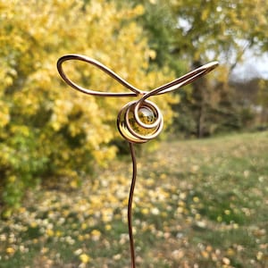 May include: A copper wire garden stake with a yellow glass orb at the top. The orb is surrounded by a copper wire spiral. The stake is in front of a blurred background of yellow and green leaves.
