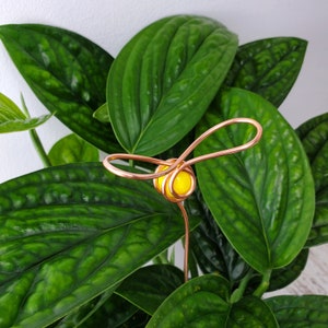 May include: A copper wire plant stake with a yellow glass bead in the center, shaped like a bee. The stake is inserted into a green potted plant.