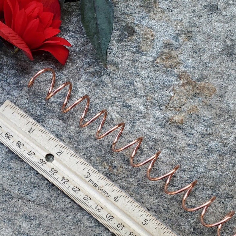 May include: A copper-colored metal coil spring, approximately 6 inches long, is lying on a gray textured surface. A ruler with the brand name "Fiskars" is partially visible in the image.