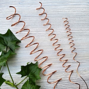 May include: Three copper wire coils of different sizes. The coils are arranged vertically on a white wooden surface. The coils are made of thin copper wire and are tightly wound.